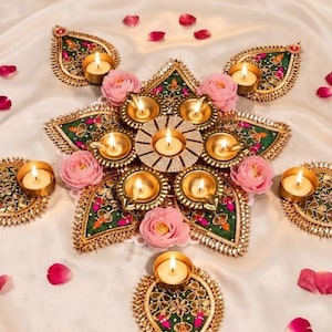 May include: A star-shaped decorative diya set featuring green fabric petals with gold embroidery and mirror accents. The arrangement includes multiple small lit tea light candles and soft pink artificial flowers, creating a symmetrical display.