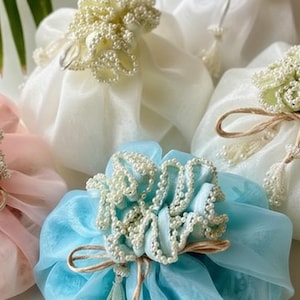 Op de afbeelding: Een verzameling decoratieve cadeauzakjes in verschillende pastelkleuren. Elke zak is gemaakt van transparante stof en versierd met een cluster van kleine, witte, parelachtige kralen. De zakken zijn vastgebonden met touw en sommige hebben kralenkwasten.