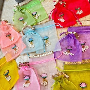 May include: A collection of colorful, sheer drawstring bags in various shades including pink, green, red, and yellow. Each bag is adorned with a lotus flower design and beaded accents, with gold trim along the top edge. The bags are arranged on a light-colored surface.