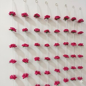 Könnte beinhalten: Dekorative Girlande aus pinkfarbenen Rosen und weißen Perlen. Jeder Strang zeigt mehrere pinkfarbene Rosen, die entlang einer Perlenkette angeordnet sind und vertikal hängen. Die Rosen haben eine tiefe rosa Farbe.