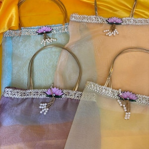 May include: Four sheer tote bags in pastel colors: blue, peach, purple, and yellow. Each bag has a gold handle, a decorative trim, and a lotus flower with pearl accents. The bags are arranged on a yellow satin surface.