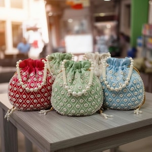Puede incluir: Una colección de cinco bolsos de terciopelo con cordón en varios colores: rojo, verde, beige y azul. Cada bolso está adornado con un delicado estampado floral y un asa de cuentas de perlas. Los bolsos se exhiben sobre una mesa de madera gris.