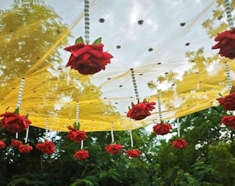 May include: A decorative ceiling installation with hanging red roses and strings of pearls. The roses are suspended from a yellow and white canopy, creating a festive atmosphere. Green foliage is visible in the background.