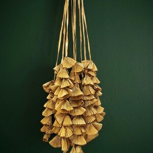May include: A collection of gold-coloured, fan-shaped tassels suspended against a dark green backdrop. The tassels are constructed from layered, textured material and hang from long, thin strands. The overall appearance is decorative and elegant.