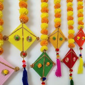Op de afbeelding: Kleurrijke handgemaakte vliegerdecoraties met slingers van gele en oranje goudsbloemen. De vliegers zijn in verschillende kleuren, waaronder roze, geel, groen, rood en blauw, met gouden accenten en kwastjes. Een feestelijke en levendige decoratie.