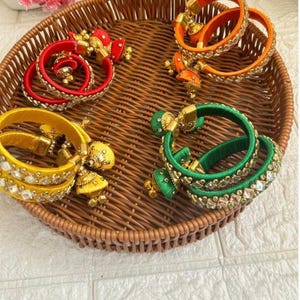 May include: A collection of colorful bangles in red, orange, yellow, and green, displayed in a woven basket. The bangles are adorned with gold-colored embellishments and sparkling accents, showcasing a traditional design.