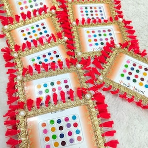 Può includere: Diversi quadri decorativi con adesivi a pois colorati. Ogni cornice ha un bordo dorato, nappe rosse e un accento di perla. Il testo "Pooja Laxmi" è visibile. Gli adesivi sono di vari colori.