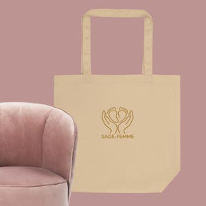 May include: Beige tote bag with a gold-colored graphic of hands cradling baby footprints, and the text "SAGE-FEMME". The bag has two handles and is made of a sturdy fabric. The background is a muted mauve color.