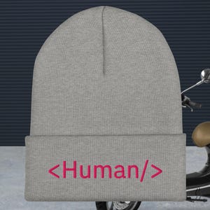 Puede incluir: Un gorro gris con el texto rosa "<Human/>". El gorro está hecho de un material de punto y tiene un dobladillo doblado. Este sombrero informal es un accesorio cómodo.