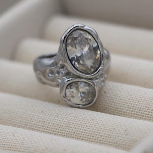Könnte beinhalten: Ein silberfarbener Ring mit zwei klaren, facettierten Steinen in ovaler und runder Form, die in ein strukturiertes, organisch wirkendes Metallband gefasst sind. Die Metalloberfläche weist ein unebenes, punktiertes Finish auf.