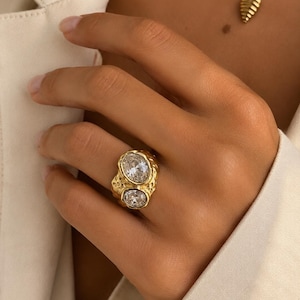 May include: A gold-toned ring featuring two clear, oval-cut stones set in a textured, organic-shaped metal band. The ring has a prominent, irregular surface finish that surrounds the two faceted stones, creating a distinct contrast between the smooth, reflective surfaces of the stones and the hammered, matte-like appearance of the gold-coloured metal setting.