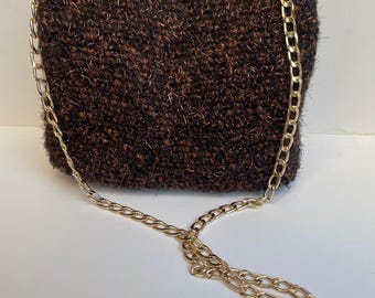 May include: A small, brown, textured handbag with a gold-colored chain strap. The bag is rectangular with rounded edges. The chain is attached to the top corners of the bag and drapes down. The bag has a soft, woven appearance.