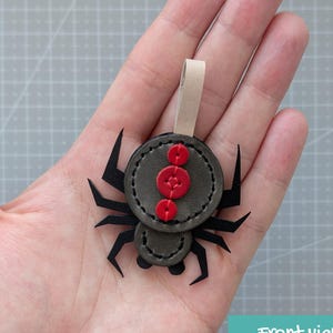 以下が含まれることがあります： ダークグレーのレザー製クモのオーナメント。8本の黒い脚と、胴体中央に縦に縫い付けられた3つの小さな赤い円形のアクセントが特徴です。上部には吊り下げ用のライトベージュのループが付いています。