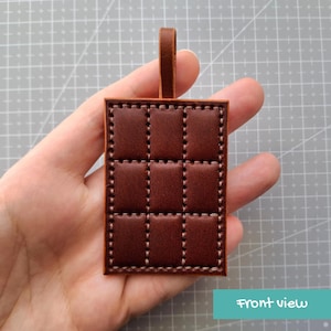 May include: A rectangular brown leather tag featuring a grid of nine padded, stitched squares and a vertical leather loop at the top. The surface has a smooth, polished finish with visible light-colored stitching outlining each individual square section.
