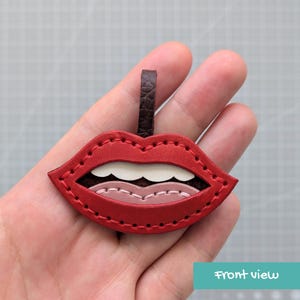 May include: A red leather keychain shaped like a pair of lips with white teeth and a pink tongue. The design features visible stitching along the edges and a dark brown leather loop at the top for attachment. The lips are layered with distinct colors.