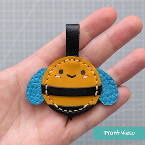 May include: A circular leather bee charm featuring a mustard yellow body with black horizontal stripes, two textured blue wings, and a black leather loop at the top. The face has two black eyes, a small black smile, and two tiny pink blush marks.