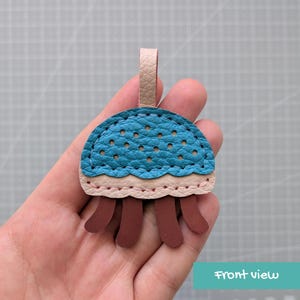 May include: A jellyfish-shaped keyring crafted from textured leather. The top dome is teal with perforated holes, attached to a light beige loop. A light beige scalloped trim sits below the dome, and four brown leather tentacles hang down.
