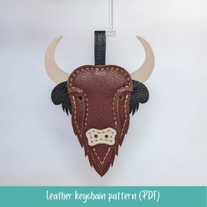 May include: A brown leather keychain in the shape of a bull's head, featuring tan horns and black accents. The keychain has a black strap and is suspended against a white background. The text "leather keychain pattern (PDF)" is at the bottom.