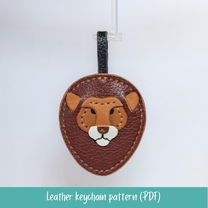 May include: A handmade leather keychain in the shape of a lion's head. The keychain features a brown oval shape with a tan mane, ears, and muzzle. The lion has a white mouth and black eyes. The text "leather keychain pattern (PDF)" is at the bottom.