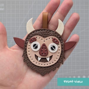May include: A brown and beige leather monster-shaped keychain with a textured surface, featuring two white horns, pointed ears, a triangular nose, and a wide mouth with two white fangs. The piece is stitched with white thread and has a brown loop.