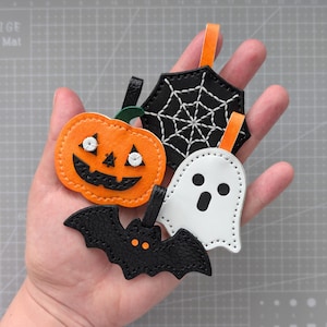 May include: A collection of Halloween-themed leather tags. The tags include an orange pumpkin, a black bat, a white ghost, and a black spiderweb. Each tag has a loop for hanging and is made with stitched details.