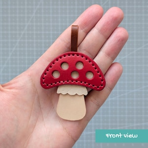 May include: A mushroom-shaped leather keychain featuring a vibrant red cap with five circular cutouts and visible black stitching. The stem is a light beige color with a scalloped edge detail, and a small brown leather loop is attached to the top.