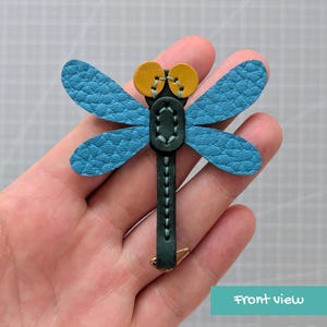 May include: A dragonfly-shaped leather accessory featuring textured blue wings, a dark green body, and two round yellow eyes. The piece is constructed with visible light-coloured stitching along the body and wings, with a small metal loop at the base.