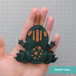 May include: A dark green leather frog-shaped keyring or charm featuring light brown leather accents on the back and head. The piece is constructed with visible light-coloured stitching along the edges and includes a small brown leather loop at the top.