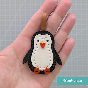 May include: A penguin-shaped leather keychain featuring a black outer body, a white belly, and a tan leather loop at the top. The design includes a small orange beak and orange feet, with visible white stitching securing the layers together.
