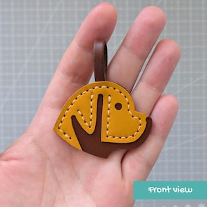 May include: A mustard yellow and dark brown leather keychain shaped like a dog head in profile. The design features a dark brown ear and muzzle section against a mustard yellow face, accented with light tan stitching and a dark brown loop.