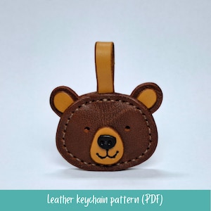 Puede incluir: Un llavero de cuero marrón con forma de oso con detalles en color beige, incluyendo las orejas y el lazo. El oso tiene una nariz negra y un contorno cosido. El texto "Leather keychain pattern (PDF)" está en la parte inferior.