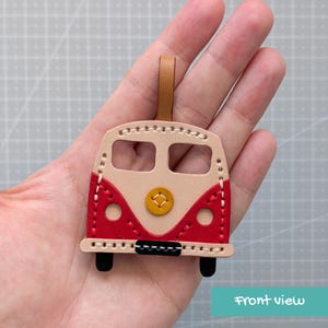 May include: A leather keychain shaped like a vintage van, featuring a beige upper section with two rectangular windows, a red lower section with two circular headlights, a central yellow circular emblem, and a small black rectangular bumper.