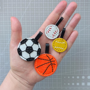 Könnte beinhalten: Eine Sammlung von Leder-Schlüsselanhängern mit Sportmotiven. Sie zeigen einen Fußball, einen Baseball, einen Basketball und einen gelben Softball. Jeder Schlüsselanhänger hat eine schwarze Schlaufe. Die Farben sind Weiß, Schwarz, Orange und Gelb.