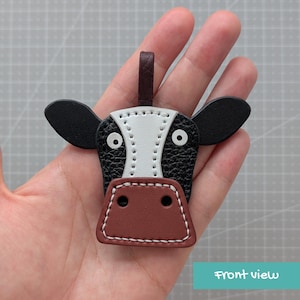 May include: A cow-shaped leather keyring featuring a black and white textured face, a brown muzzle, and two black ears. The design includes a small dark brown leather loop at the top for attachment, with white stitching detailing the facial features.