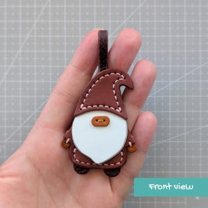 May include: A brown leather gnome keyring with a white beard and a brown hat. The gnome has a button nose and is stitched with white thread. The keyring has a brown loop for attaching to keys or a bag.