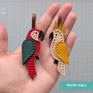 May include: Two leather parrot-shaped keychains with brown leather loops. One parrot is red with a dark green wing and black beak. The other is light beige with a yellow wing, yellow crest, and black beak, both featuring visible stitching.