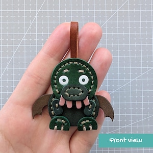 Op de afbeelding: Een handgemaakte groene leren monster sleutelhanger met bruine accenten. De sleutelhanger heeft een gestikt ontwerp met witte ogen, een roze mond en kleine bruine vleugels. Een bruine leren lus is bevestigd om op te hangen.