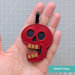 May include: A red leather skull-shaped keychain with a black leather backing and a black loop at the top. The skull features two dark eye sockets and two rows of light tan leather teeth accents, all secured with visible stitching along the edges.