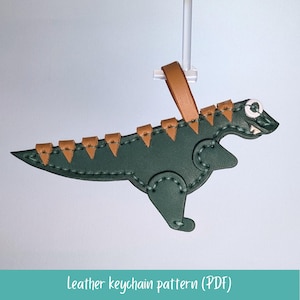 以下が含まれることがあります： 茶色のアクセントが付いた緑色の革製恐竜キーホルダー。キーホルダーには取り付け用のループがあり、ステッチのデザインが施されています。「Leather keychain pattern (PDF)」というテキストが下にあります。