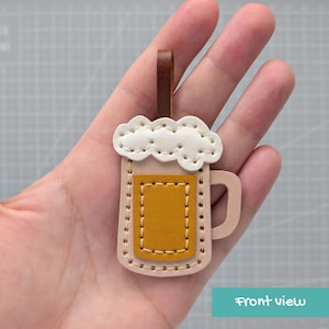 以下が含まれることがあります： ベージュ、マスタードイエロー、ブラウンの層状レザーで作られたビールジョッキ型のキーホルダー。クリーム色の泡のトップ、マスタードイエローの中央パネル、上部には取り付け用のブラウンのレザーループが特徴的なデザインです。