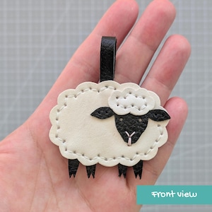 May include: A sheep-shaped keychain made of cream and black leather. The body is cream with white stitching, while the head, ears, and legs are black. A black leather loop extends from the top. The sheep has a small pink stitched nose.