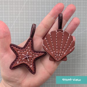 May include: Two brown leather keychains: a starfish and a seashell. Both have white stitching and a dark brown loop. The starfish has a five-point design. The seashell keychain has a fan-like pattern.
