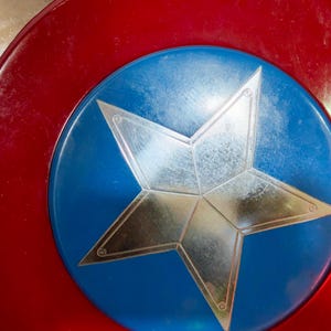 May include: A shield featuring a red border, a blue central disc, and a prominent silver star. The shield's surface shows signs of wear. The design is similar to that of a superhero's emblem, with a classic, iconic look.