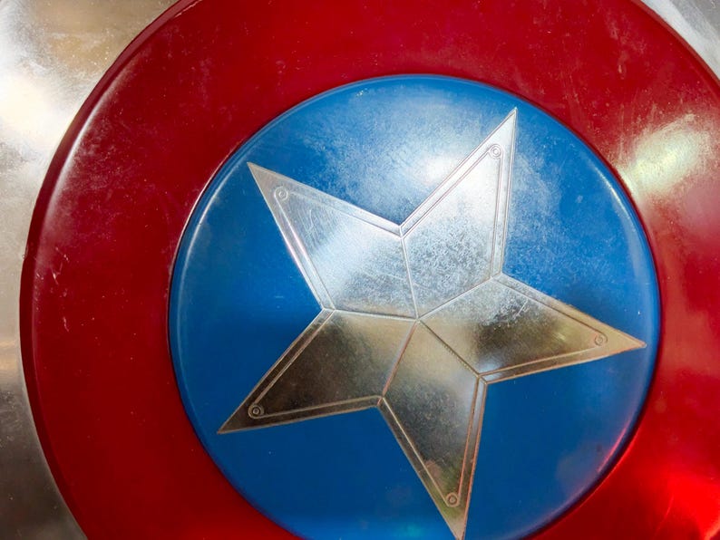 May include: A shield featuring a red border, a blue central disc, and a prominent silver star. The shield's surface shows signs of wear. The design is similar to that of a superhero's emblem, with a classic, iconic look.