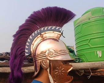Puede incluir: Un casco romano de color cobre con una pluma morada. El casco presenta un diseño de sol en la mejilla y espirales decorativas. La pluma es larga y fluida, añadiendo un toque dramático a este tocado histórico.