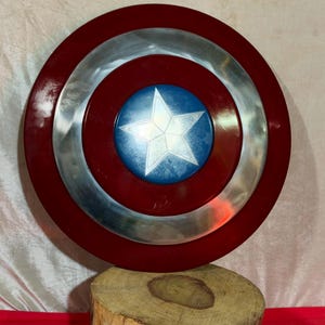 May include: A circular shield featuring a red border, a silver central ring, and a blue central disc with a white star. The shield is displayed on a wooden base. This is a replica of Captain America's shield.