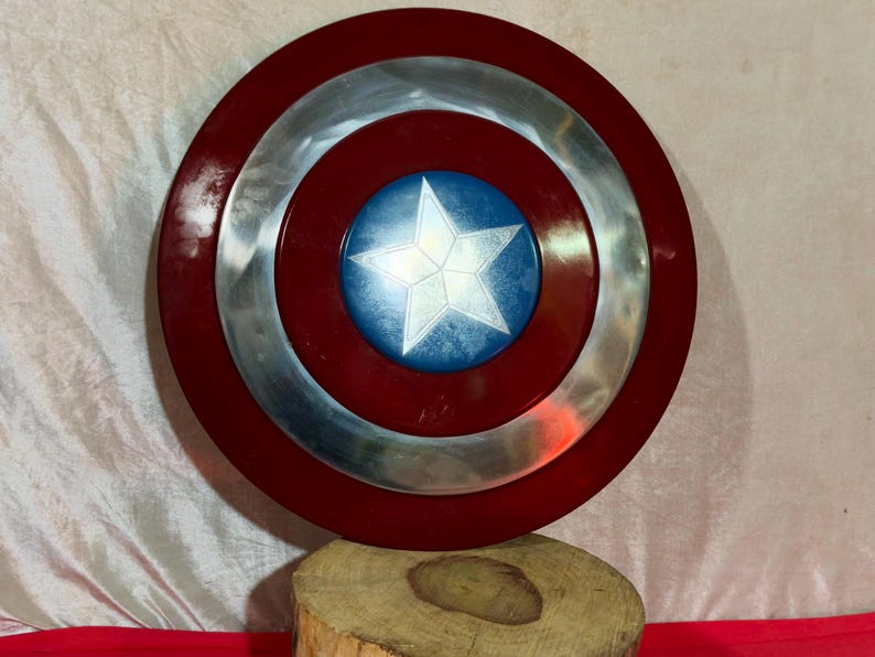 May include: A circular shield featuring a red border, a silver central ring, and a blue central disc with a white star. The shield is displayed on a wooden base. This is a replica of Captain America's shield.
