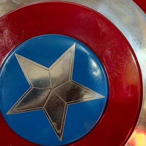 May include: Close-up of a Captain America shield. The shield features a red, white, and blue colour scheme. A silver star is centred on a blue circle, surrounded by red and silver concentric rings. The shield has a metallic appearance.