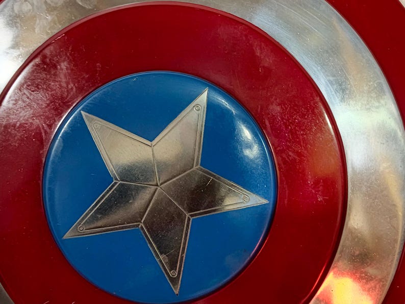 May include: Close-up of a Captain America shield. The shield features a red, white, and blue colour scheme. A silver star is centred on a blue circle, surrounded by red and silver concentric rings. The shield has a metallic appearance.
