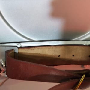 May include: Close-up of a brown leather strap with a gold-coloured buckle, attached to a white object with silver metal hardware. The leather strap has a natural edge and is secured with rivets.